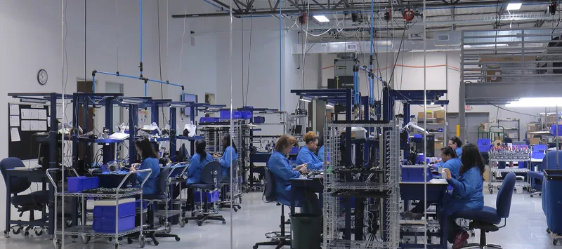 Electronic assembly floor with blue-smocked technicians at wire-rack workstations