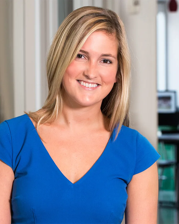 professional headshot smiling blonde woman blue v-neck top blurred indoor background