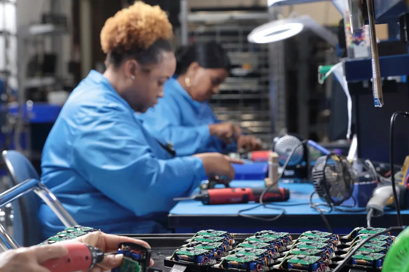 Electronic assembly technicians in blue uniforms soldering circuit boards at workbench