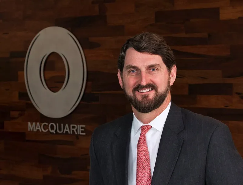 Corporate headshot executive in dark suit smiling before branded wood-panel backdrop